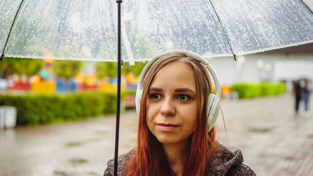 Młoda piękna kobieta w słuchawkach słucha muzyki stojącej z przezroczystym parasolem w deszczu na ulicy miasta