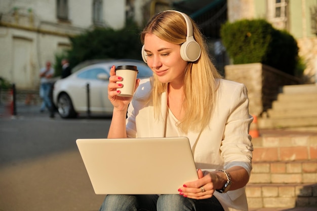 Młoda piękna kobieta w słuchawkach z laptopem pijąca kawę w papierowym kubku