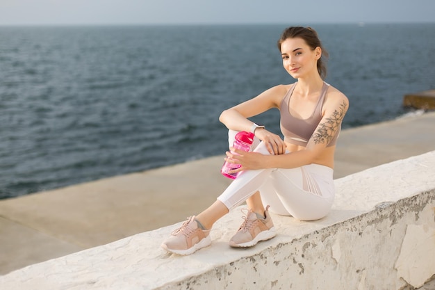 Młoda piękna kobieta w sportowym top i białych legginsach, trzymając w ręku różową butelkę sportową, jednocześnie szczęśliwie patrząc w aparacie z widokiem na morze na tle