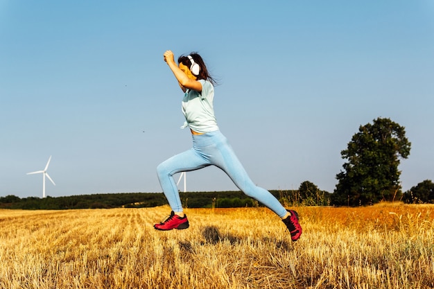 Młoda piękna kobieta w średnim wieku skoki w polu zbóż w hełmach i odzieży sportowej. zdrowy tryb życia. wiatraki na horyzoncie. energie odnawialne. Troska o planetę