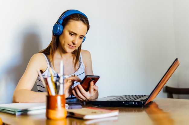 Młoda piękna kobieta w średnim wieku w domu siedzi przy stole z komputerem i patrzy na telefon komórkowy ze słuchawkami. koncepcja telepraca.technologia