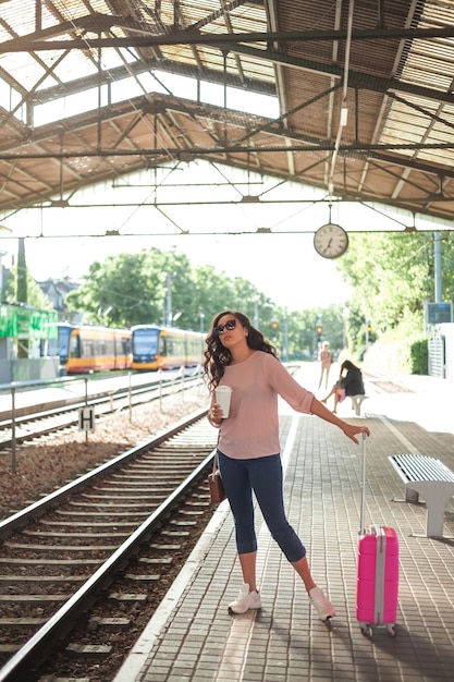 Młoda piękna kobieta w wieku 3040 lat z różową małą walizką czeka na pociąg na stacji Kręcone modelki w swobodnych ubraniach rozmawia przez telefon pije kawę na wynos Podróż