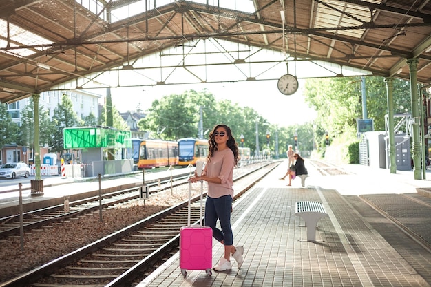Młoda piękna kobieta w wieku 3040 lat z różową małą walizką czeka na pociąg na stacji Kręcone modelki w swobodnych ubraniach rozmawia przez telefon pije kawę na wynos Podróż