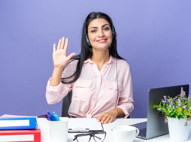 Młoda piękna kobieta w zwykłych ubraniach nosi zestaw słuchawkowy z mikrofonem, uśmiechając się, pokazując numer pięć, siedząc przy stole z laptopem nad niebieską ścianą, pracując w biurze