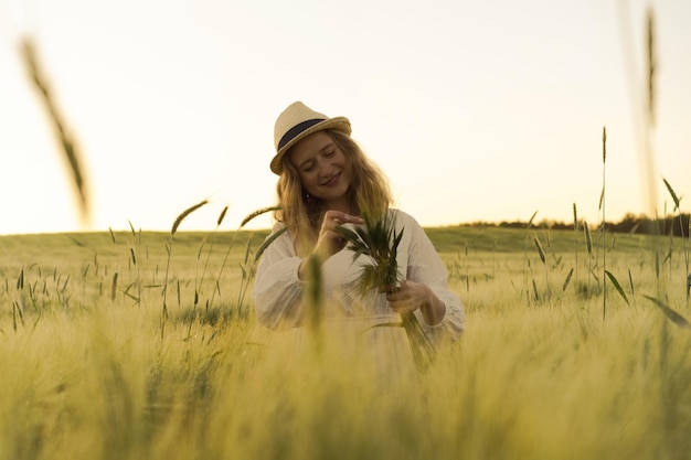 młoda piękna kobieta z blond długimi włosami w białej sukni w słomkowym kapeluszu zbiera kwiaty na polu pszenicy. Latające włosy w słońcu, lato. Czas dla marzycieli, złoty zachód słońca.