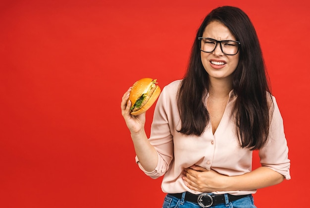 Młoda piękna kobieta z bólem brzucha na czerwonym tle Jedzenie niezdrowego fast food burgera lub kanapki