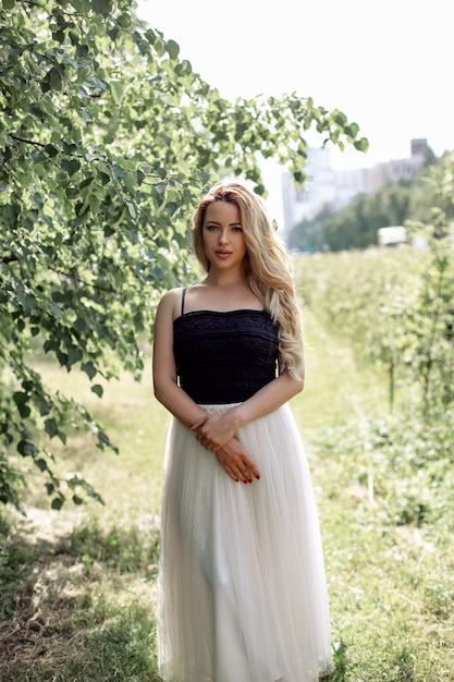 młoda piękna kobieta z długimi kręconymi blond włosami i makijażem w koronkowej spódnicy spacerująca w letnim parku