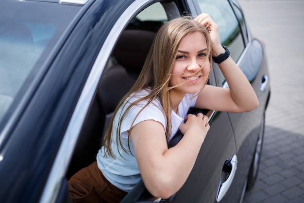 Młoda piękna kobieta z długimi włosami siedzi w czarnym samochodzie na parkingu. Ładna dziewczyna w ubraniu. Podróż samochodem