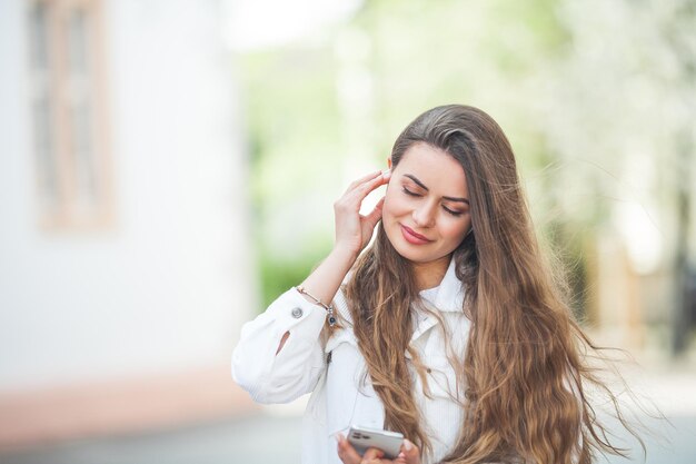 Młoda piękna kobieta z długimi włosami w wygodnych, lekkich, codziennych ubraniach słucha muzyki za pomocą słuchawek bluetooth spaceruje po centrum europejskiego miasta Śliczna modelka robi zakupy Wiosna