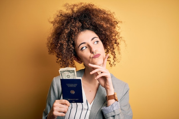 Młoda piękna kobieta z kręconymi włosami i piercingiem, trzymająca niemiecki paszport i poważną twarz dolarów, myśląca o bardzo zdezorientowanym pomyśle