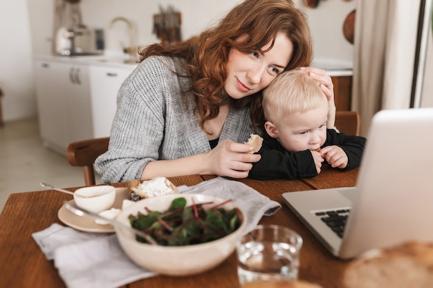 Młoda piękna kobieta z rudymi włosami w swetrze z dzianiny przytulająca swojego małego synka siedzącego przy stole z jedzeniem i oglądając bajki na laptopie