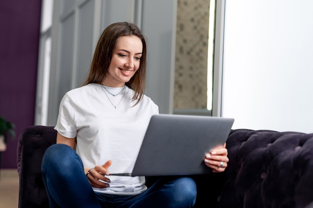 Młoda piękna kobieta za pomocą laptopa w domu. Czarna sofa. Uśmiechnięta młoda kobieta z laptopem.