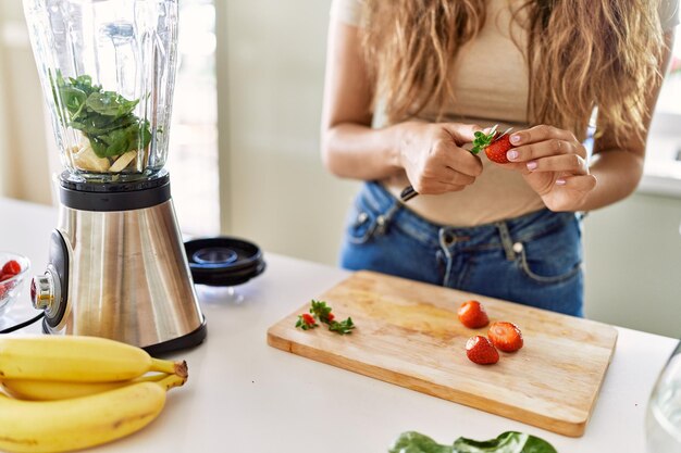 Młoda piękna latynoska kobieta przygotowuje warzywny smoothie z blenderem tnącym truskawki w kuchni