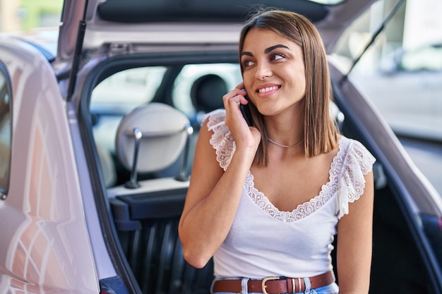 Młoda piękna latynoska kobieta rozmawia na smartfonie siedząc na bagażniku samochodu na ulicy