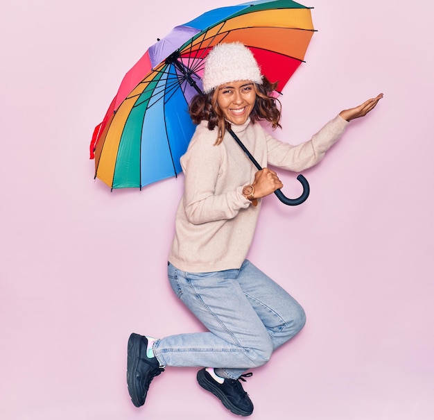 Zdjęcie młoda piękna latynoska trzymająca parasol uśmiechnięty szczęśliwy skacząc z uśmiechem na twarzy na odizolowanym różowym tle