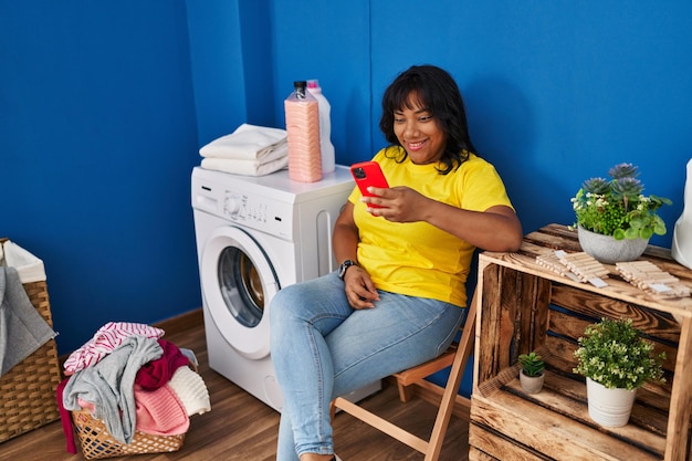 Młoda piękna Latynoska za pomocą smartfona czeka na pralkę w pralni