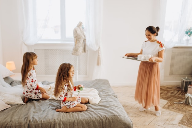 Młoda piękna mama przynosi córkom kakao z piankami i ciasteczkami siedzącymi na łóżku w pełnym światła przytulnym pokoju.