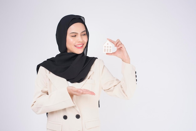 Młoda piękna muzułmańska kobieta w garniturze, trzymająca mały modelowy dom na białym tle studio