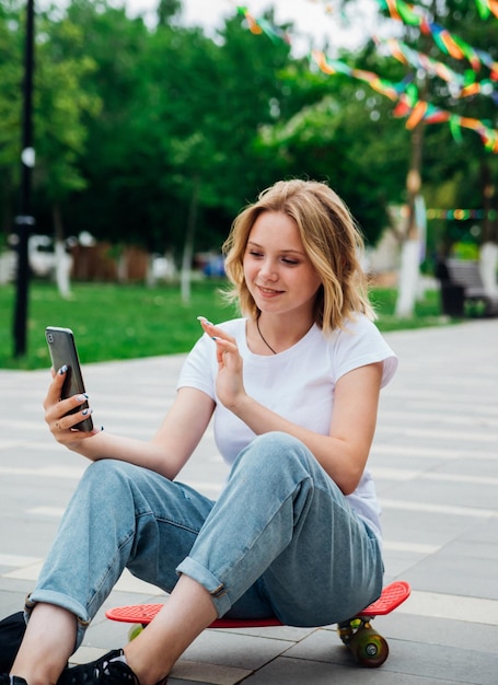 Młoda piękna nastolatka siedzi na deskorolce w piękny letni dzień w parku Dziewczyna rozmawia przez wideo przez telefon komórkowy na świeżym powietrzu Czat z przyjaciółmi