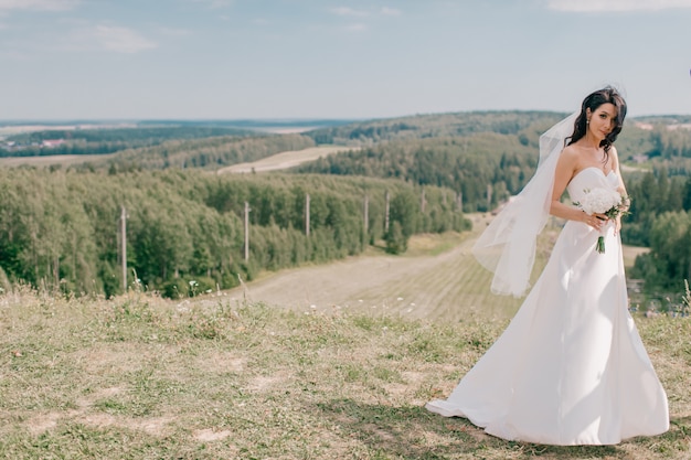 Młoda piękna panna młoda w białej ślubnej sukni pozuje na wzgórzu przy naturą.