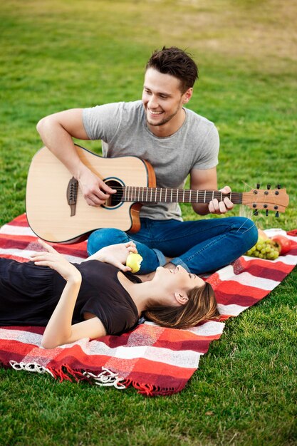 Młoda piękna para uśmiechnięta, odpoczynek, relaks na pikniku w parku. Mężczyzna gra na gitarze.