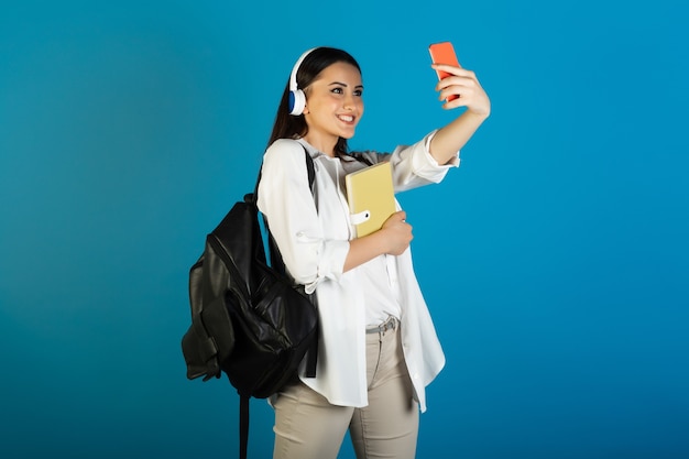 Młoda piękna studentka z plecakiem i słuchawkami, trzymając żółty notatnik i sprawia, że selfie na niebiesko