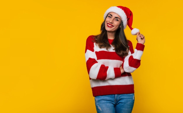 Młoda piękna szczęśliwa dziewczyna w Santa hat i zimowym swetrze pozuje na białym tle na żółtym tle w czasie świąt Bożego Narodzenia