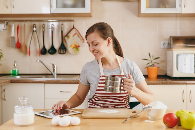 Młoda piękna szczęśliwa kobieta przesiewa mąkę przez sito i szuka w kuchni przepisu na ciasta w tabletce. Gotowanie w domu. Przygotuj jedzenie.