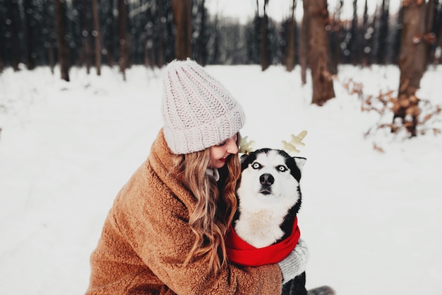 Młoda Piękna Szczęśliwa Uśmiechnięta Kobieta W Futrze Przytulanie Psa Husky Ubrany W Czerwony Szalik świąteczny I świąteczne Rogi Jelenia. Zdjęcie Portretowe Dziewczyny I Psa Na Białym śniegu W Zimowym Lesie. Szczęśliwego Nowego Roku