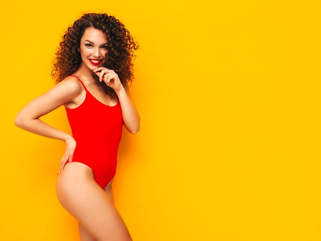 Młoda piękna uśmiechnięta kobieta pozuje w pobliżu żółtej ściany w studioSeksowna modelka w czerwonym stroju kąpielowymPozytywna kobieta z lokami fryzuraSzczęśliwa i wesoła