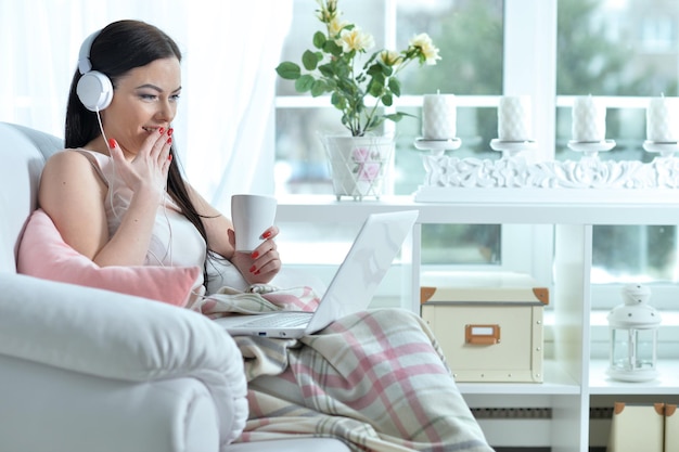 Młoda piękna uśmiechnięta kobieta siedzi przy fotelu i korzysta z laptopa