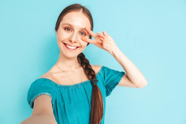 Młoda piękna uśmiechnięta kobieta w modnych letnich dżinsach kombinezonach Seksowna beztroska kobieta z fryzurą na ogon pozuje przy ścianie w studio Pozytywny model zabawy Wesoły i szczęśliwy Robienie zdjęć selfie