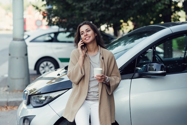 Młoda Piękna Uśmiechnięta Kobieta Z Filiżanką Kawy Mówi Na Smartfonie W Pobliżu Swojego Pojazdu Elektrycznego