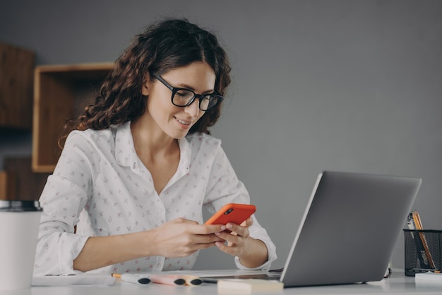 Młoda piękna włoska bizneswoman korzystająca z nowoczesnego smartfona podczas pracy zdalnej na laptopie