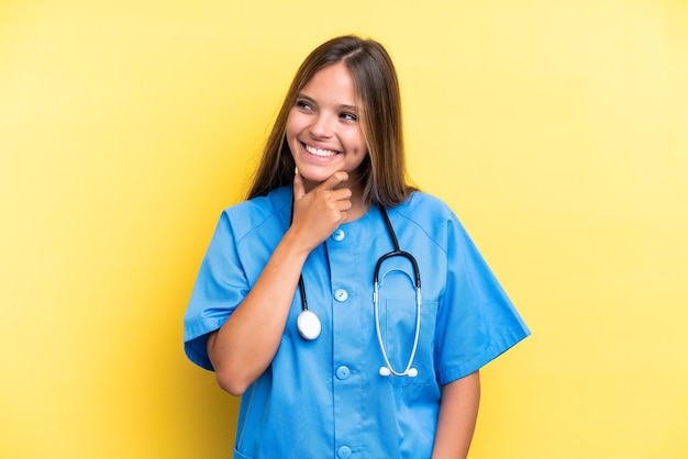 Młoda pielęgniarka kaukaska kobieta odizolowana na żółtym tle, patrząc w bok i uśmiechnięta
