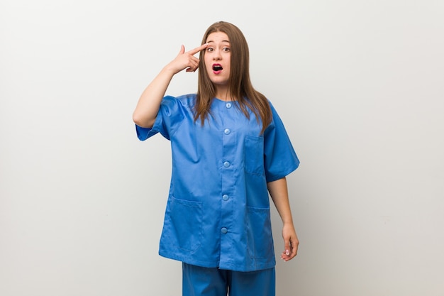 Młoda pielęgniarka kobieta na białej ścianie pokazuje gest rozczarowania palcem wskazującym.