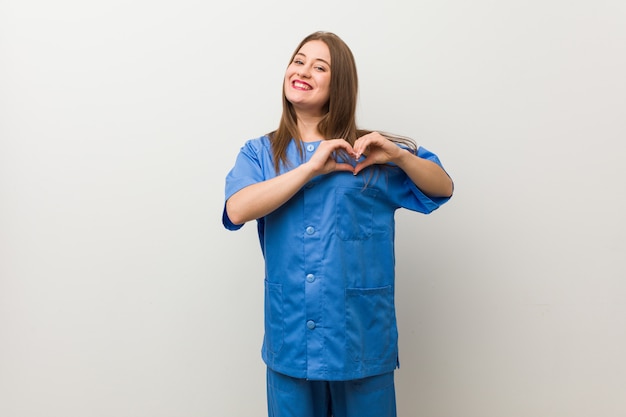 Młoda Pielęgniarka Kobieta Na Białej ścianie, Uśmiechając Się I Pokazując Kształt Serca Rękami.
