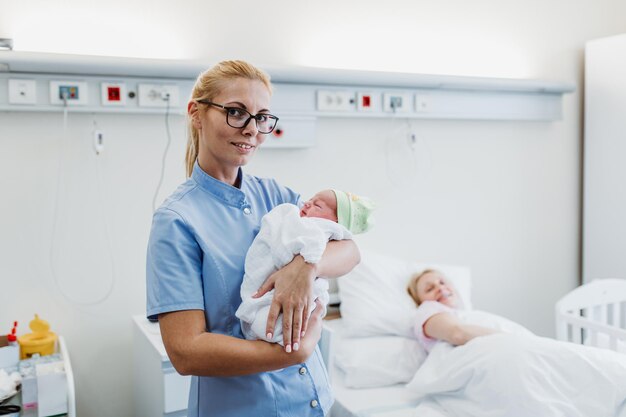 Młoda Pielęgniarka Stojąca Na Oddziale Położniczym I Trzymająca W Ramionach Noworodka. Koncepcja Po Urodzeniu.