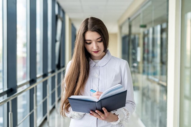 Młoda pielęgniarka w błękitnym mundurze pozuje w szpitalnym korytarzu z stetoskopem i magazynem