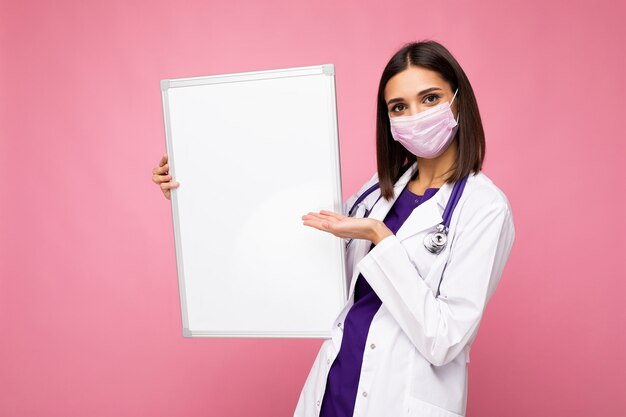 Młoda pielęgniarka w ochronnej masce na twarz i białym fartuchu medycznym trzymająca pustą tablicę magnetyczną