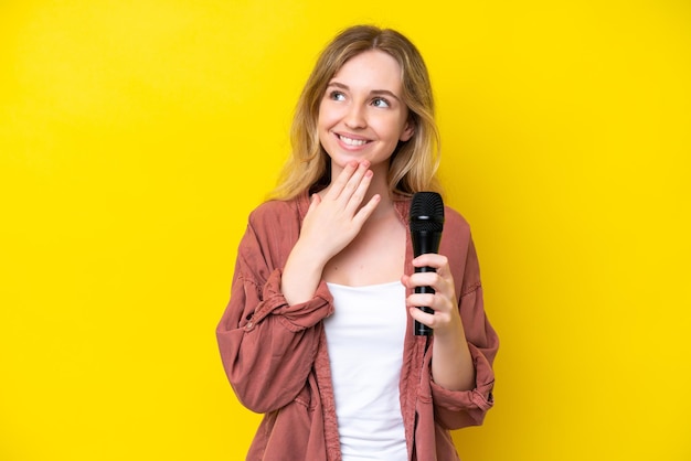 Młoda piosenkarka kaukaska kobieta podnosi mikrofon odizolowany na żółtym tle, patrząc w górę, uśmiechając się