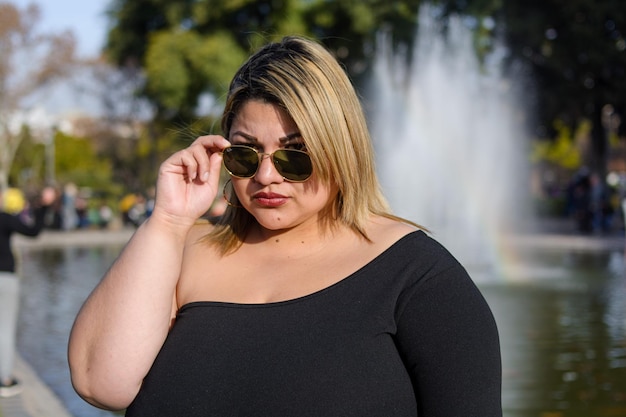 młoda plus size modna kobieta pochodzenia łacińskiego argentyńskiego w parku pozowanie z okularami przeciwsłonecznymi