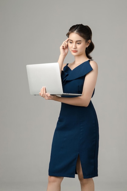 Młoda poważna azjatycka kobieta biznesu trzymająca laptopa i myśląca na szarym tle
