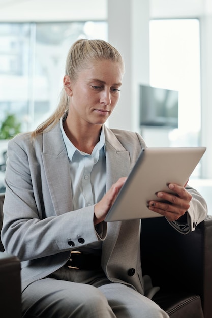 Młoda poważna blond bizneswoman w szarym eleganckim garniturze za pomocą tabletu