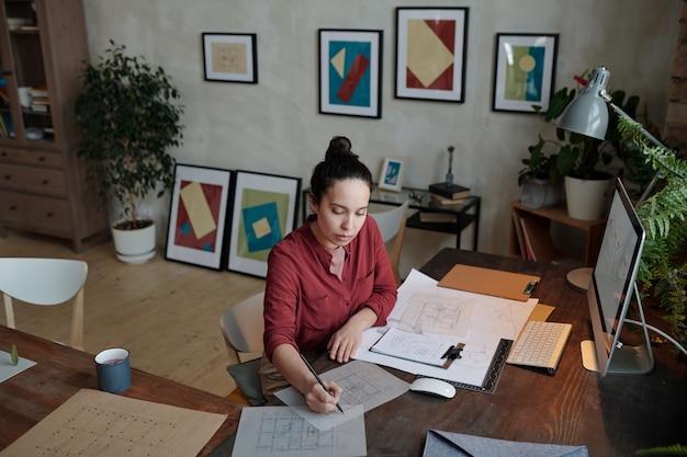 Młoda poważna kobieta architekt z ołówkiem rysunek szkic nowej konstrukcji, siedząc przy stole przed monitorem komputera