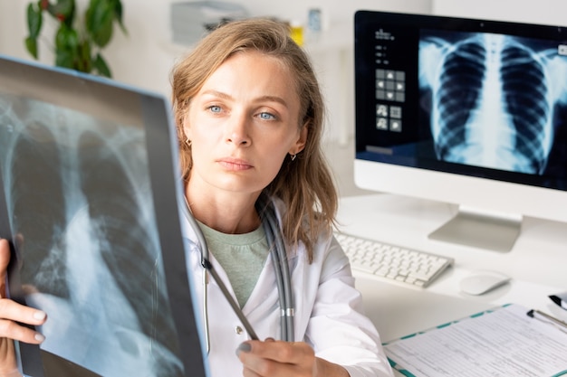 Młoda poważna kobieta radiolog w białym płaszczu, wskazując na zdjęcie rentgenowskie płuc pacjenta, siedząc przed miejscem pracy z monitorem komputera