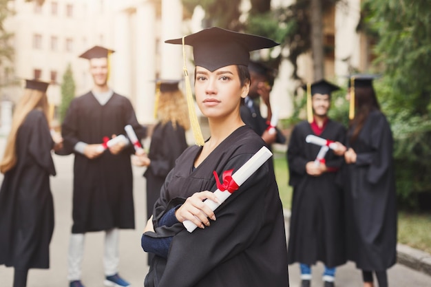 Młoda poważna kobieta w dniu ukończenia studiów na uniwersytecie, stojąc z wieloetniczną grupą przyjaciół. Koncepcja edukacji, kwalifikacji i sukni.