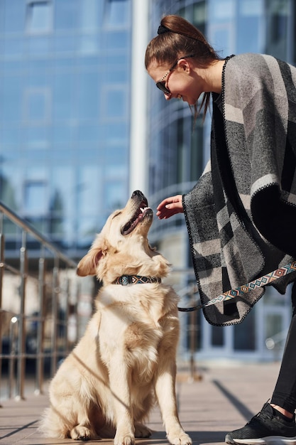 Młoda pozytywna kobieta bawi się z psem podczas spaceru na świeżym powietrzu w pobliżu budynku biznesowego