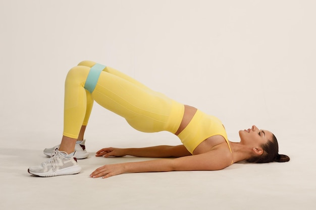 Młoda pozytywna uśmiechnięta kobieta z ekspanderem na nogi i ręce w żółtym stroju sportowym fitness studio na białym tle
