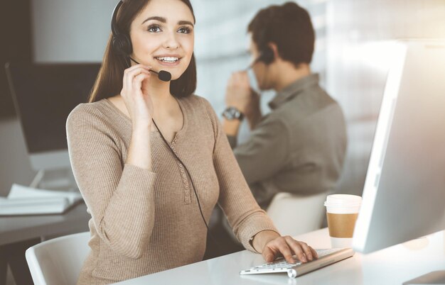 Młoda przyjazna dziewczyna w zestawach słuchawkowych rozmawia z klientem firmy, siedząc przy biurku w słonecznym biurze. Operatorzy call center w pracy.